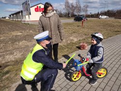 policjant drogówki kuca przy dziecku na rowerku i podaje opaskę odblaskową, obok stoi matka dziecka